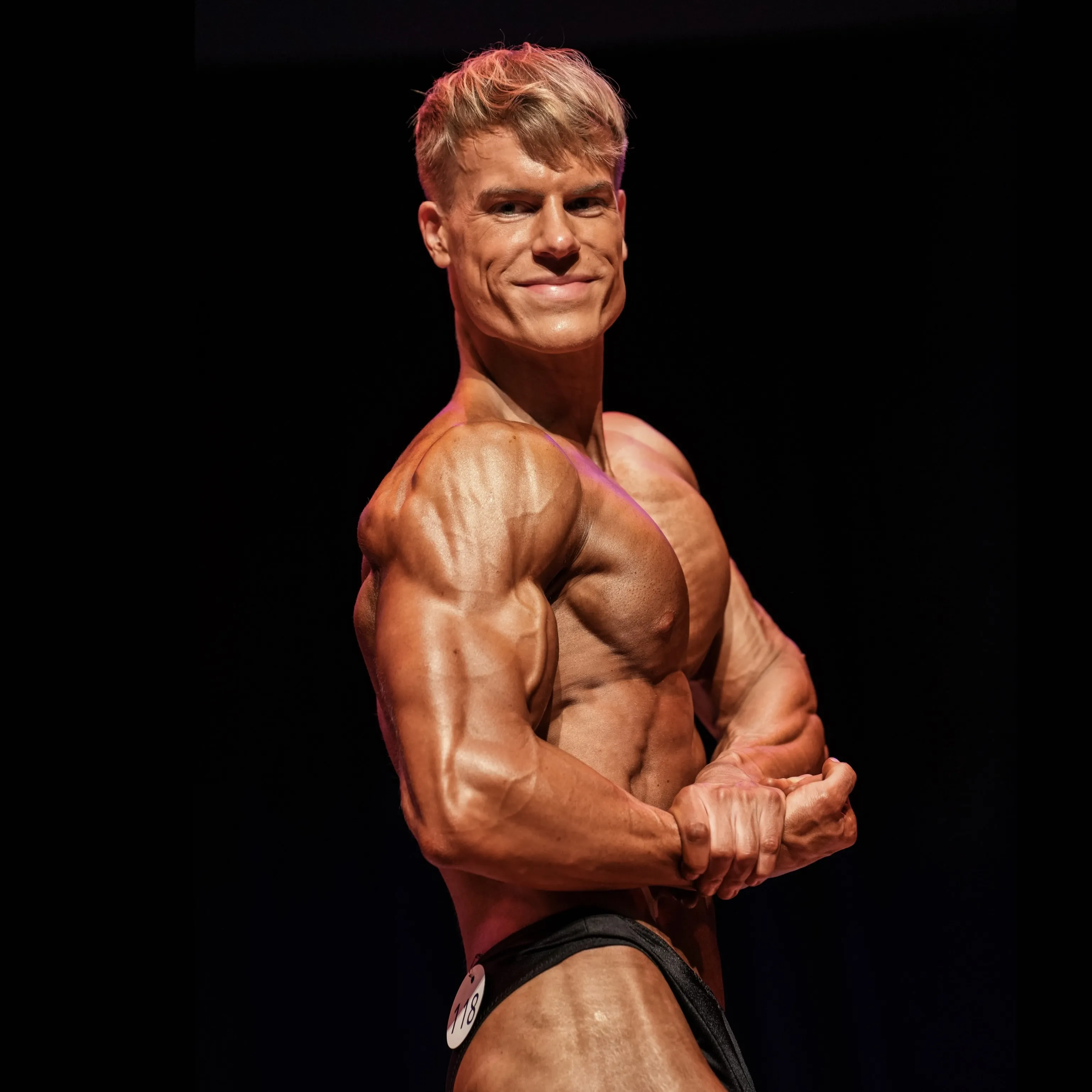Bodybuilder posing on stage in a side chest position, showcasing well-defined muscles under stage lighting.